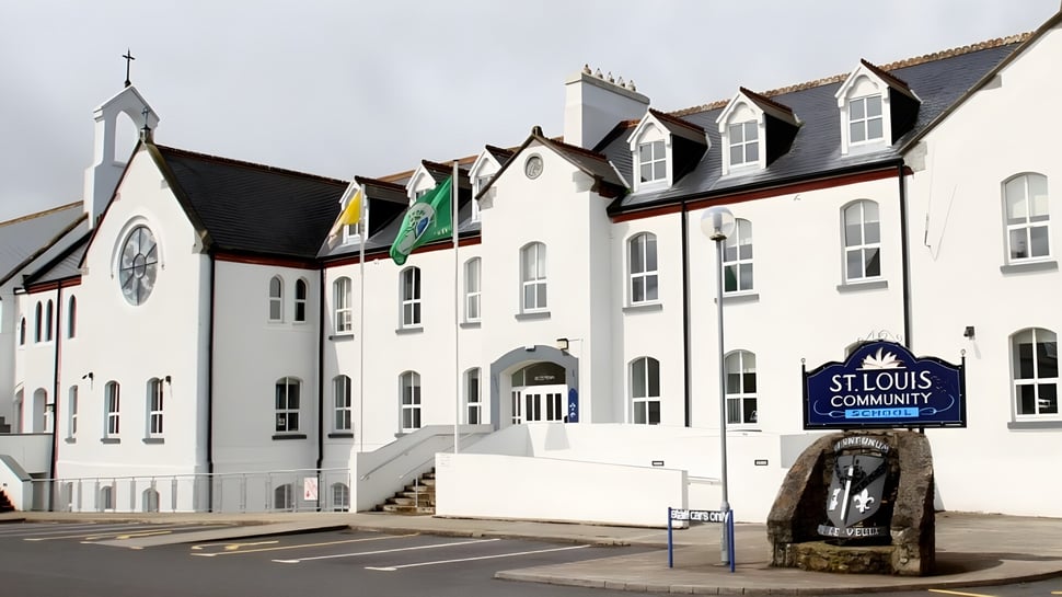El edificio blanco de varios pisos con una torre similar a un campanario y banderas en el terreno de la St. Louis Community School con un monumento de piedra en primer plano.