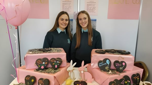 Dos alumnas de la St. Louis Secondary School están frente a un fondo rosa con dos grandes pasteles decorados con corazones al frente.