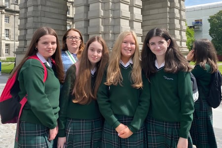 Un grupo de estudiantes de la St. Louis Secondary School en uniforme escolar está frente a un edificio histórico con un área verde al fondo.