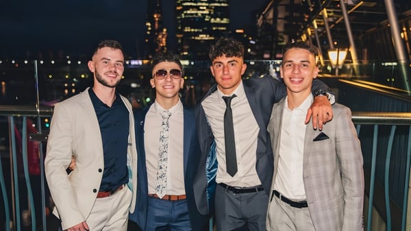 Cuatro estudiantes de St. Macartan's College posan juntos frente a un paisaje urbano nocturno con rascacielos iluminados.