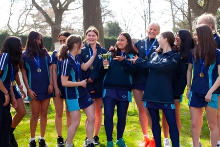 Un grupo de alumnas con uniformes oscuros y azules uniformes está de pie en un campo con árboles al fondo en el terreno de la St. Margaret’s School.