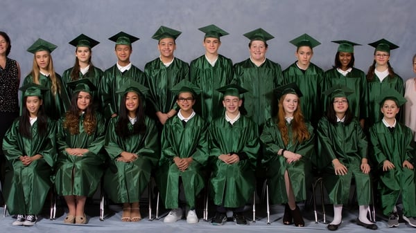 Un grupo de graduados de la St. Mark High School está de pie en togas y birretes verdes frente a un fondo gris.