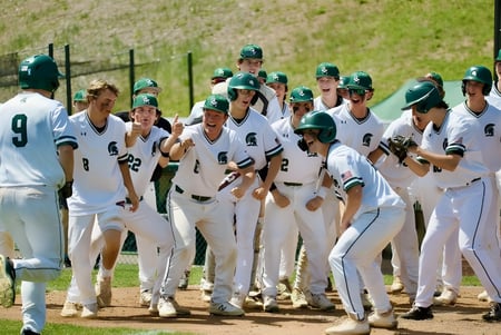 Un grupo de jugadores de béisbol en uniformes blancos con cascos verdes en el campo de béisbol de la St. Mark High School.
