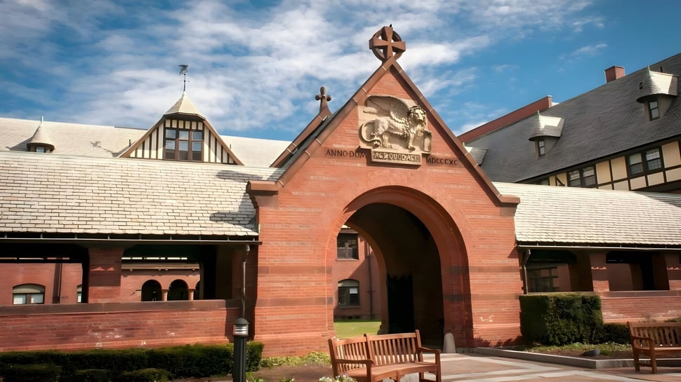 El característico edificio de ladrillo de la St. Mark's School con techo puntiagudo y portal de arco está rodeado de vegetación verde.
