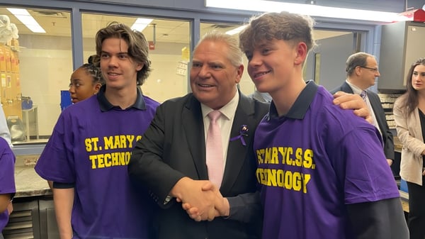 Tres personas en camisetas moradas saludan a un hombre mayor en traje en la St. Mary Catholic Secondary School.