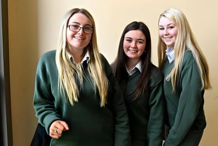 Tres alumnas en uniformes escolares verdes están sonriendo juntas en el pasillo de St. Mary's Holy Faith Killester.