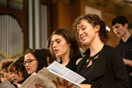 Un grupo de estudiantes de la St. Mary’s Music School canta juntos en un escenario con fondo de madera.