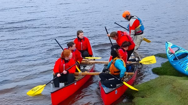 Un grupo de estudiantes de St. Mary’s Secondary lleva chalecos salvavidas rojos y se encuentra con kayaks en un cuerpo de agua cerca de la orilla.