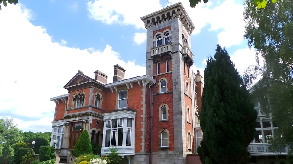 El imponente edificio de ladrillo con alto torre en el campus de St. Michael's College está rodeado de vegetación verde bajo un cielo azul.