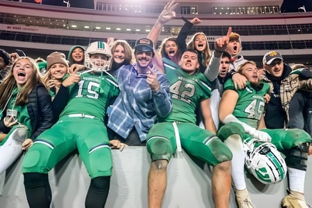Un grupo de estudiantes de la St. Mother Teresa High School anima en el estadio con camisetas y chaquetas verdes.