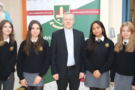 Un grupo de alumnas y alumnos del St. Patricks College en uniforme escolar está con un hombre mayor frente a una pancarta verde y roja.