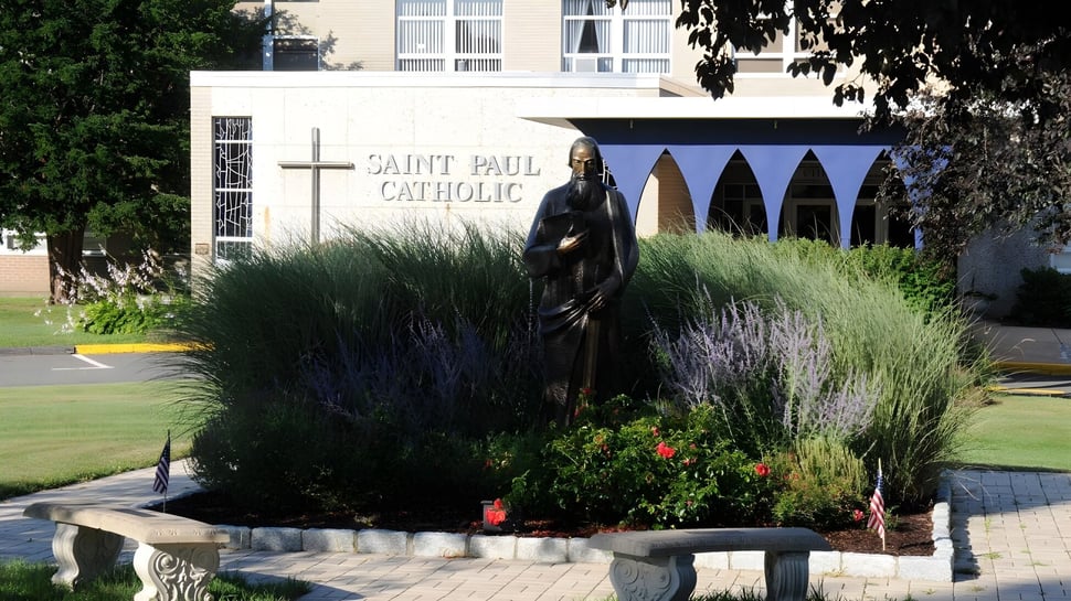 Una estatua de una figura religiosa se encuentra en el jardín bien cuidado en el terreno de la St. Paul Catholic High School con la iglesia católica de fondo.