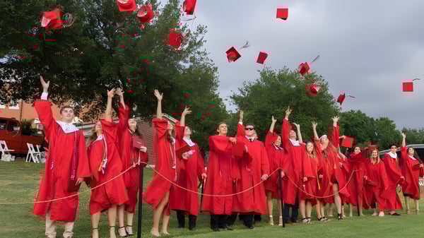 Graduados de la St. Paul High School en túnicas rojas están de pie con los brazos en alto durante la ceremonia de graduación frente a unos árboles.