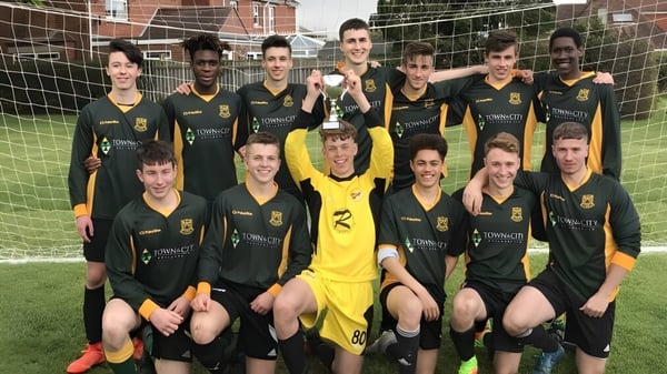 Un grupo de estudiantes de la St. Peter’s High School posan con un trofeo de fútbol en el campo frente a un edificio de ladrillo.