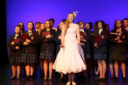 Una mujer con un vestido blanco está frente a un grupo de alumnas en el escenario de St. Peter's School Cambridge.