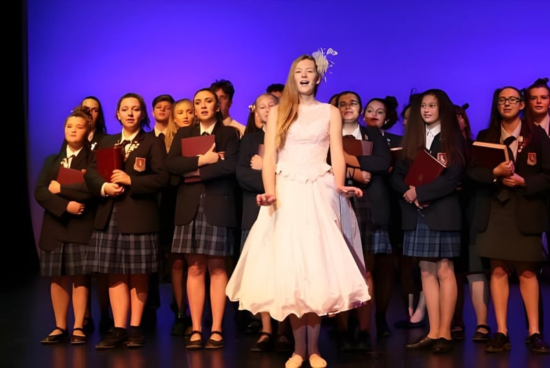Una mujer con un vestido blanco está frente a un grupo de alumnas en el escenario de St. Peter's School Cambridge.