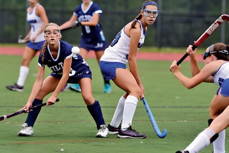 Estudiantes de la St. Raphaela’s Secondary School juegan un partido de hockey en un campo con césped verde y árboles al fondo.