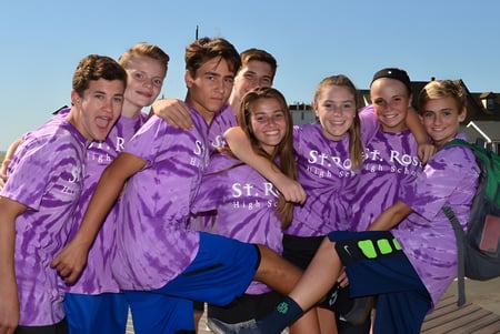 Un grupo de alumnas y alumnos de la St. Rose High School lleva camisetas de batik moradas y está de pie juntos en una plataforma de madera.