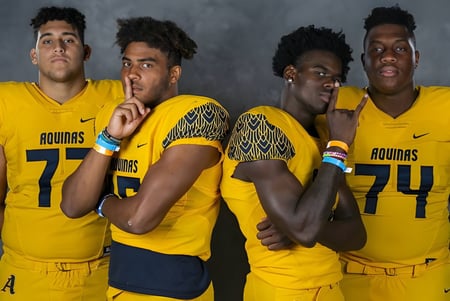 Cuatro estudiantes de la St. Thomas Aquinas High School llevan camisetas amarillas de Aguias y posan juntos frente a un fondo oscuro.