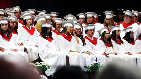Las graduadas y graduados de la St. Thomas Aquinas High School están sentados en togas blancas con estolas rojas juntos en un escenario.