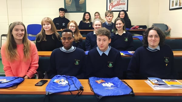 Estudiantes de la St. Tiernan’s Community School están sentados en uniformes escolares en mesas con materiales escolares.
