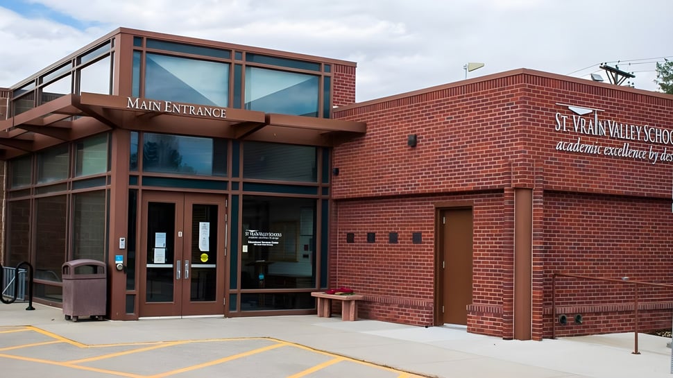 El edificio de ladrillo con la entrada principal de St. Vrain Valley Schools se encuentra bajo un cielo nublado y está rodeado de un área pavimentada.