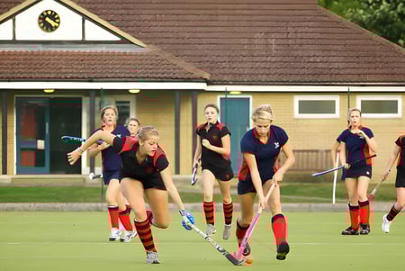 Un grupo de alumnas de Stamford Endowed Schools juega al hockey sobre césped en un campo frente a un edificio tradicional.