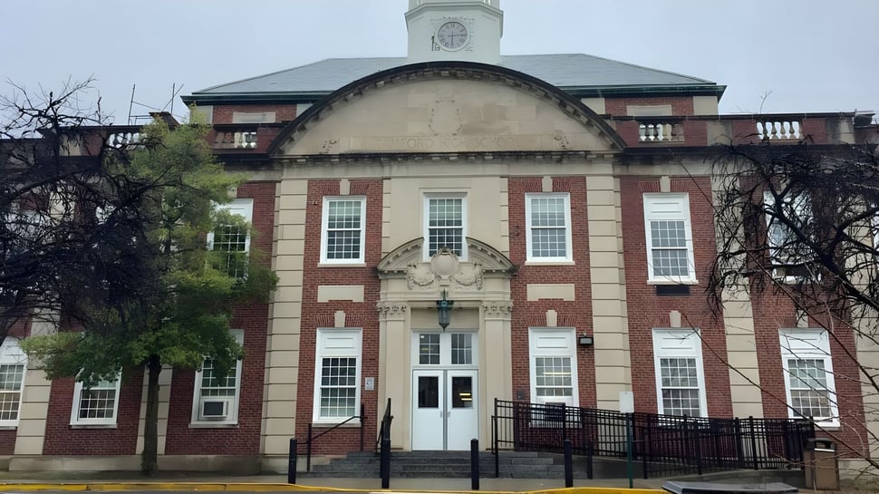 El edificio de ladrillo con torre del reloj de Stamford High School está rodeado de árboles desnudos y una cerca de hierro forjado.