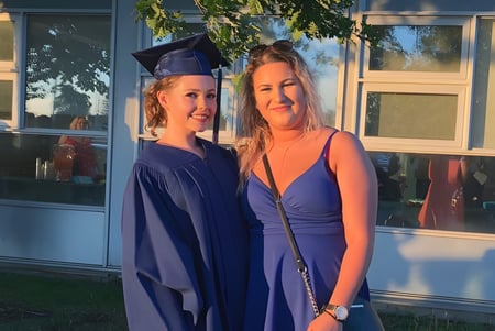 Dos alumnas, una con una toga de graduación, están frente al edificio del Stayner Collegiate Institute.