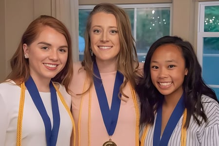 Tres graduadas de la Stearns High School en ropa de graduación sostienen medallas frente a una ventana con vista al verde.