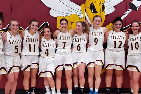 Un grupo de estudiantes de Stellys Secondary School está en camisetas de baloncesto blancas frente a una pared roja con una gran figura de caricatura.