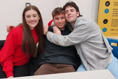 Tres alumnas y alumnos se abrazan y sonríen en el campus de la Stepaside Educate Together Secondary School frente a muros de información.