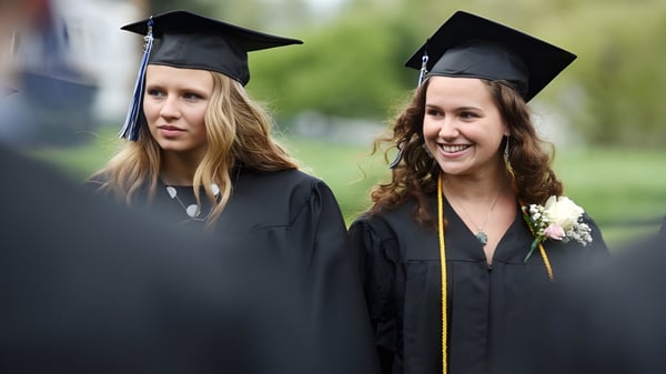 Dos graduadas de la Stillwater Christian School están juntas en sus togas de graduación frente a un fondo verde.