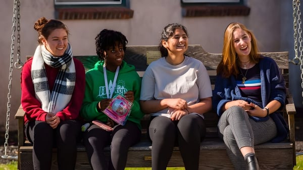 Cuatro estudiantes están sentadas en un banco de madera y conversan en el campus de la Stoneleigh-Burnham School.