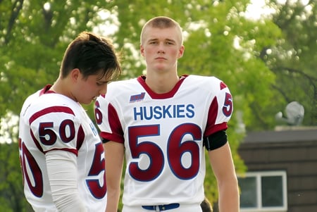 Dos estudiantes en camisetas de Huskies están en el área verde del Collège Sturgeon Heights Collegiate frente a unos árboles.