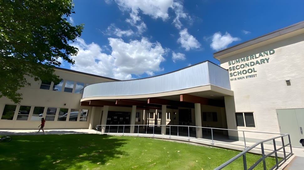 El moderno edificio de dos pisos de la Summerland Secondary School con entrada curva y césped en primer plano bajo un cielo azul.