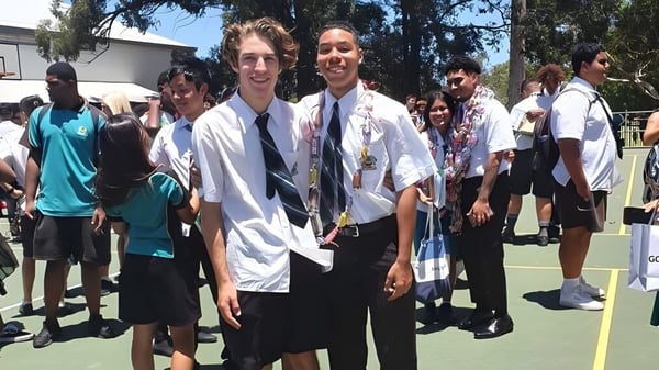 Un grupo de estudiantes está junto afuera en el campus de la Sunnybank State High School con árboles y edificios de fondo.