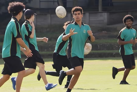 Estudiantes de la Sunnybank State High School juegan al rugby en un campo deportivo con el edificio escolar de fondo.