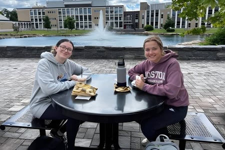 Dos estudiantes de la SUNY Plattsburgh están sentadas en una mesa frente a una fuente y comen un bocadillo en el campus.