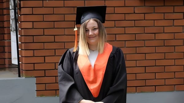 Una estudiante en ropa de graduación está frente a una pared de ladrillo en el campus del Superior Collegiate & Vocational Institute.
