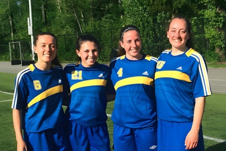 Cuatro estudiantes de la Sutherland Secondary School están juntas en uniformes deportivos azules y amarillos en un campo cubierto de hierba.