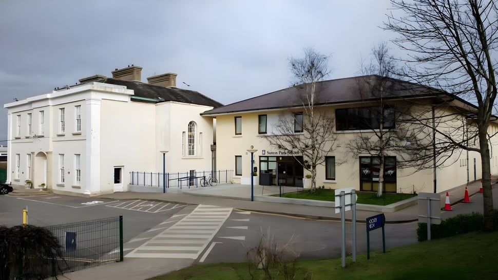 El edificio blanco de varios pisos con techo inclinado de la Sutton Park School en Dublín está rodeado de árboles desnudos.
