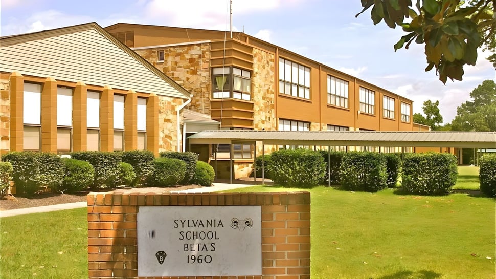 El edificio de dos pisos de la Sylvania High School, con revestimiento de ladrillo y paneles laterales, está rodeado de césped bien cuidado y arbustos.