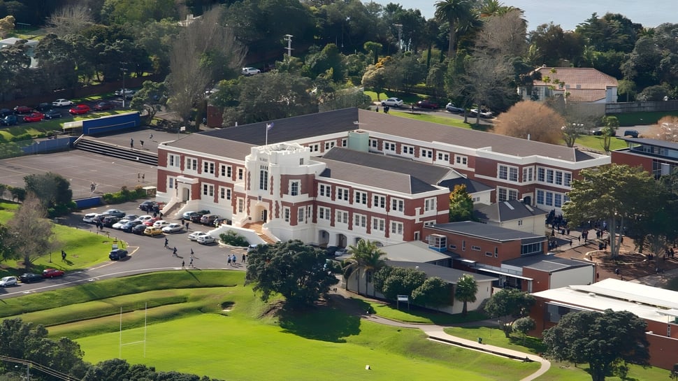 El edificio de ladrillo de varios pisos de la Takapuna Grammar School se encuentra en una colina cubierta de hierba rodeado de árboles.