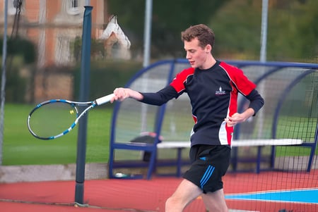 Un estudiante de Taunton School International juega al tenis en una colorida cancha al aire libre con edificios y árboles de fondo.