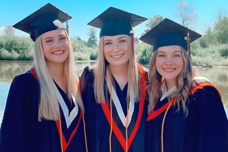 Tres estudiantes de la Terry Fox Secondary School posan con vestimenta de graduación frente a un lago con un bosque de fondo.