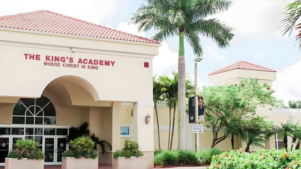 El gran edificio de The King's Academy con techo de tejas rojas está rodeado de palmeras y vegetación.