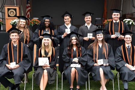 Graduadas y graduados de The Linsly School en vestimenta académica están frente a un edificio con una bandera americana.