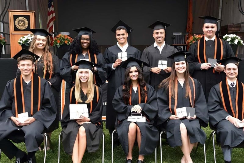 Graduadas y graduados de The Linsly School en vestimenta académica están frente a un edificio con una bandera americana.