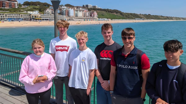 Estudiantes de The Oratory School están en un muelle mirando hacia una ciudad costera con edificios coloridos y mar azul.